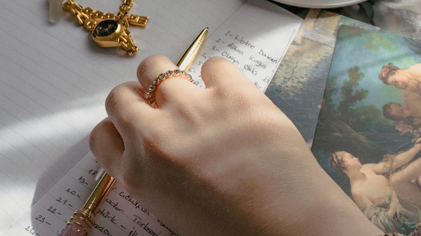 A jewelled hand writing a list beside gold jewellery and postcards