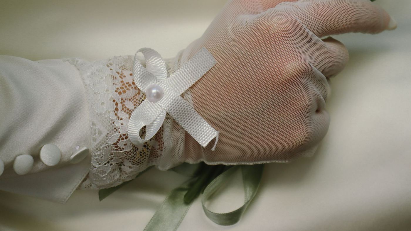 A bride's lace-gloved hand holding ribbons and a pearl bow